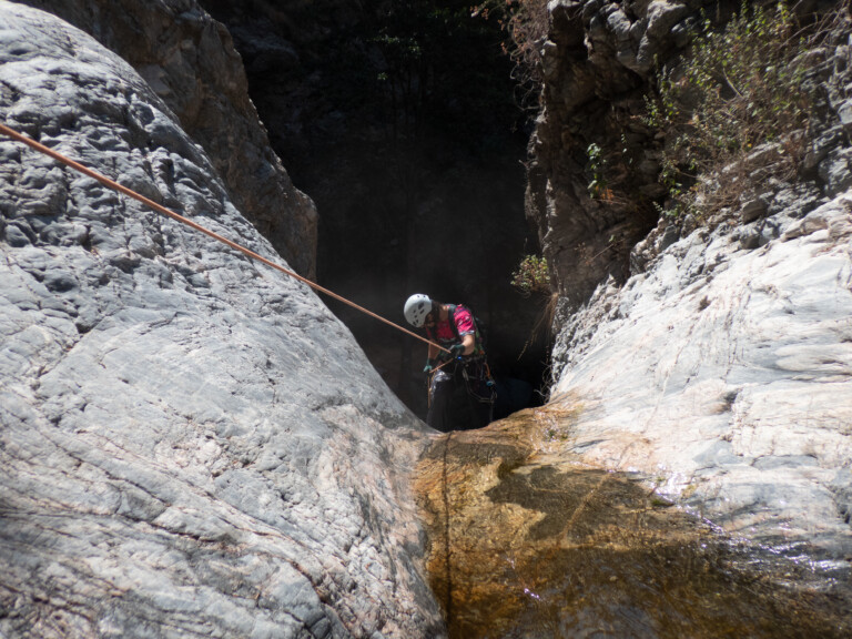 A person equipped with a white helmet, gloves, and harness is rappelling down a steep rocky canyon wall using a rope. The rocky surface is light gray with some water flowing down, and there is dense vegetation visible at the top of the canyon. The scene is illuminated by natural sunlight.