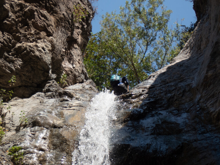 A person wearing a backpack and outdoor gear is rappelling down a steep rocky waterfall in Eaton Canyon. The scene shows rugged rock formations on both sides, water splashing down the rocks, and green trees against a clear blue sky in the background.