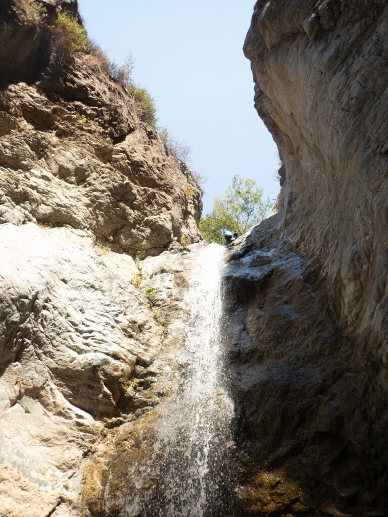 View looking up at a waterfall flowing down rocky canyon walls in Eaton Canyon. A person wearing canyoneering gear is seen descending near the top of the waterfall under a clear blue sky.