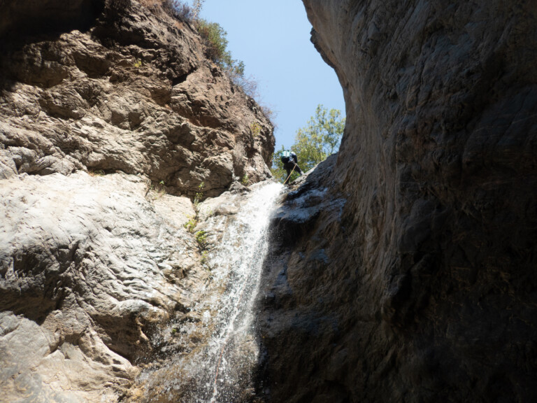 A person wearing a backpack and helmet rappelling down a waterfall in a narrow rocky canyon with steep cliffs on both sides and a clear blue sky above, captured during a canyoneering adventure in Eaton Canyon.