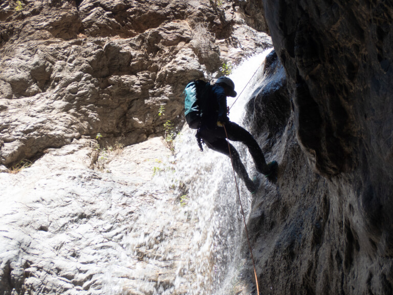 A person equipped with a helmet, backpack, and climbing gear rappels down a waterfall in a rocky canyon. Water cascades beside them as they descend the steep rock face, surrounded by rugged cliffs and sparse vegetation.