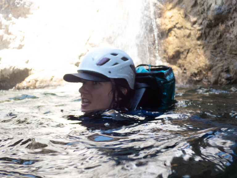 Close-up of a person wearing a white helmet and a black cap swimming in water near a rocky waterfall in Eaton Canyon. The person has a focused expression and is partially submerged in the water, with a backpack visible on their back.