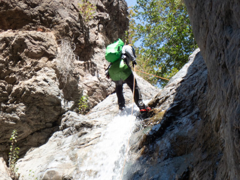 A person with a green backpack and helmet rappels down a rocky waterfall in Eaton Canyon, surrounded by rugged rock formations and green trees under a clear blue sky.