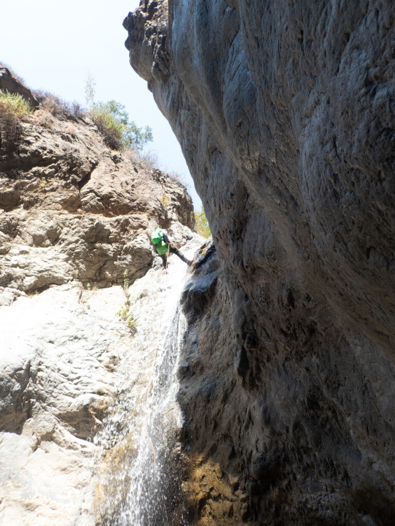A person with a green backpack rappels down a waterfall in a steep rocky canyon under bright daylight, surrounded by rugged rock formations and sparse vegetation.