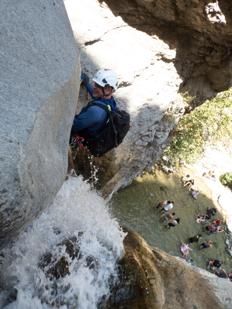 A man in a white helmet and blue jacket is canyoneering, rappelling down a rocky waterfall with water splashing around him. Below, several people are gathered in a shallow pool of water surrounded by rocks and greenery, enjoying the natural setting at Eaton Canyon.