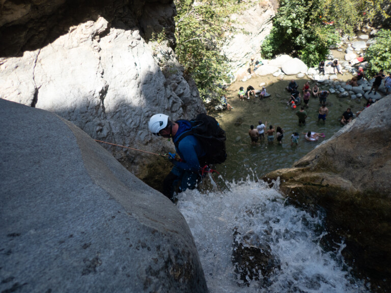 A person equipped with a white helmet, blue jacket, and backpack is rappelling down a steep rocky waterfall in Eaton Canyon. Below, a group of people stand and sit in shallow water surrounded by rocks and greenery, enjoying the outdoor environment under bright sunlight.