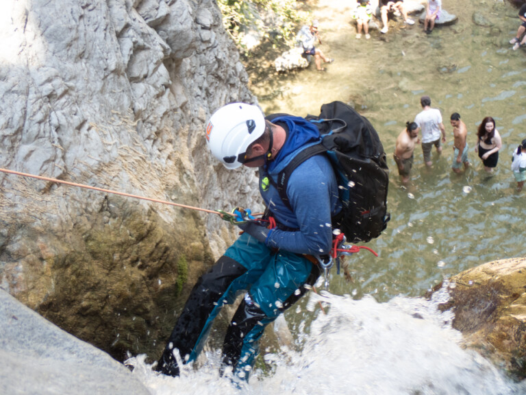 A person in blue outdoor gear and a white helmet is rappelling down a rocky waterfall using ropes and harnesses. Water splashes around their legs as they descend. Below, several people stand and sit in shallow water, observing the activity. The scene is set in a natural canyon environment with rocky walls and clear water.