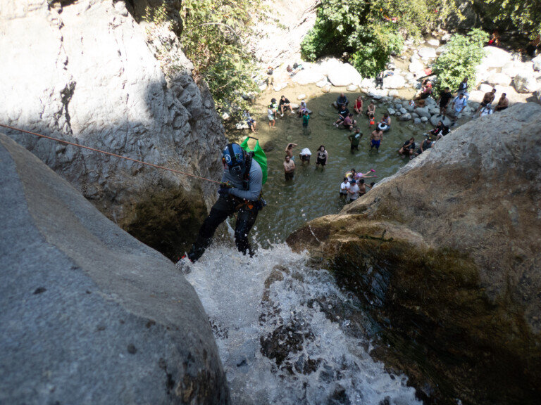 A person wearing a helmet and harness rappelling down a rocky waterfall into a shallow pool where a crowd of people stand and sit on rocks, surrounded by greenery and sunlight in Eaton Canyon.