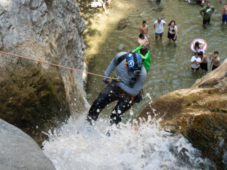 A person in a blue helmet and gray long-sleeve shirt rappelling down a rocky waterfall with water splashing around. Below, several people stand in shallow water watching, some in swimwear and others in casual clothes. The scene is set in a rocky canyon with natural water pools.
