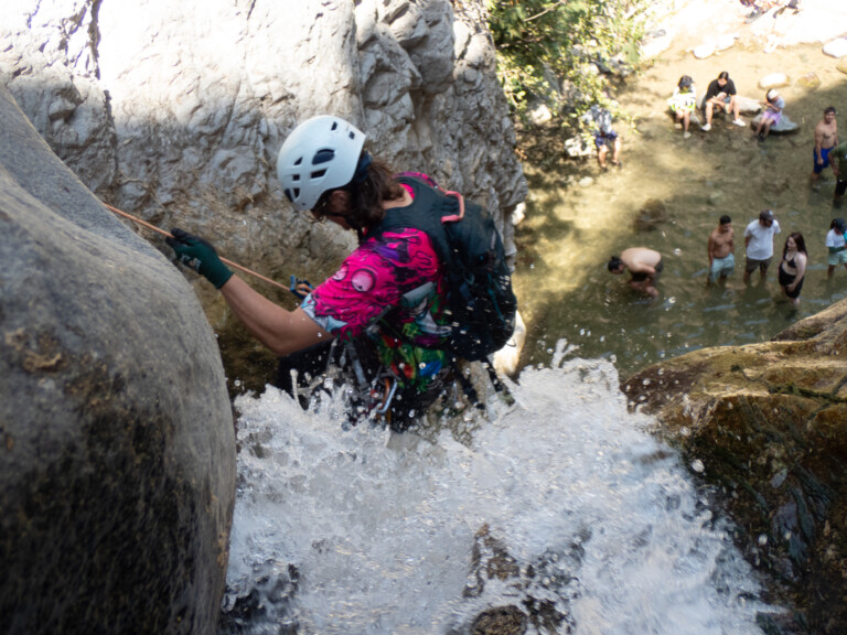 A person in a white helmet and vibrant pink and purple shirt is rappelling down a rocky waterfall with water splashing around. Below, several people stand and sit in shallow water near rocks, observing the activity. The scene is set in a natural canyon environment with rocky cliffs and some greenery.