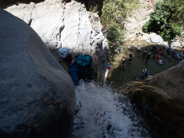 View from above of a person wearing a white helmet and teal backpack rappelling down a rocky waterfall into a shallow pool where many people are sitting and standing in the water surrounded by rocks and greenery.