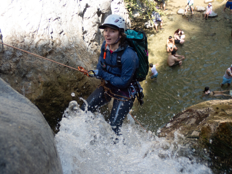 A person in a white helmet, blue jacket, and climbing harness rappels down a rocky waterfall with water splashing around. Below, several people are sitting and standing in shallow water surrounded by rocks and trees, enjoying the outdoor setting.