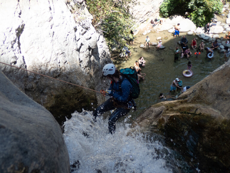 A person equipped with a white helmet, blue jacket, and green backpack is rappelling down a rocky waterfall with splashing water. Below, a group of people, including children and adults, are gathered in a shallow pool surrounded by rocks and greenery, enjoying the water and sunshine.