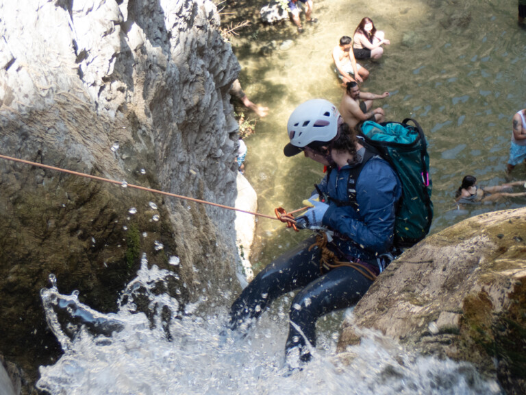 A person in a white helmet, blue jacket, and black pants is rappelling down a rocky waterfall with water splashing around. Below, several people in swimwear sit and stand in shallow water surrounded by rocks and natural scenery.