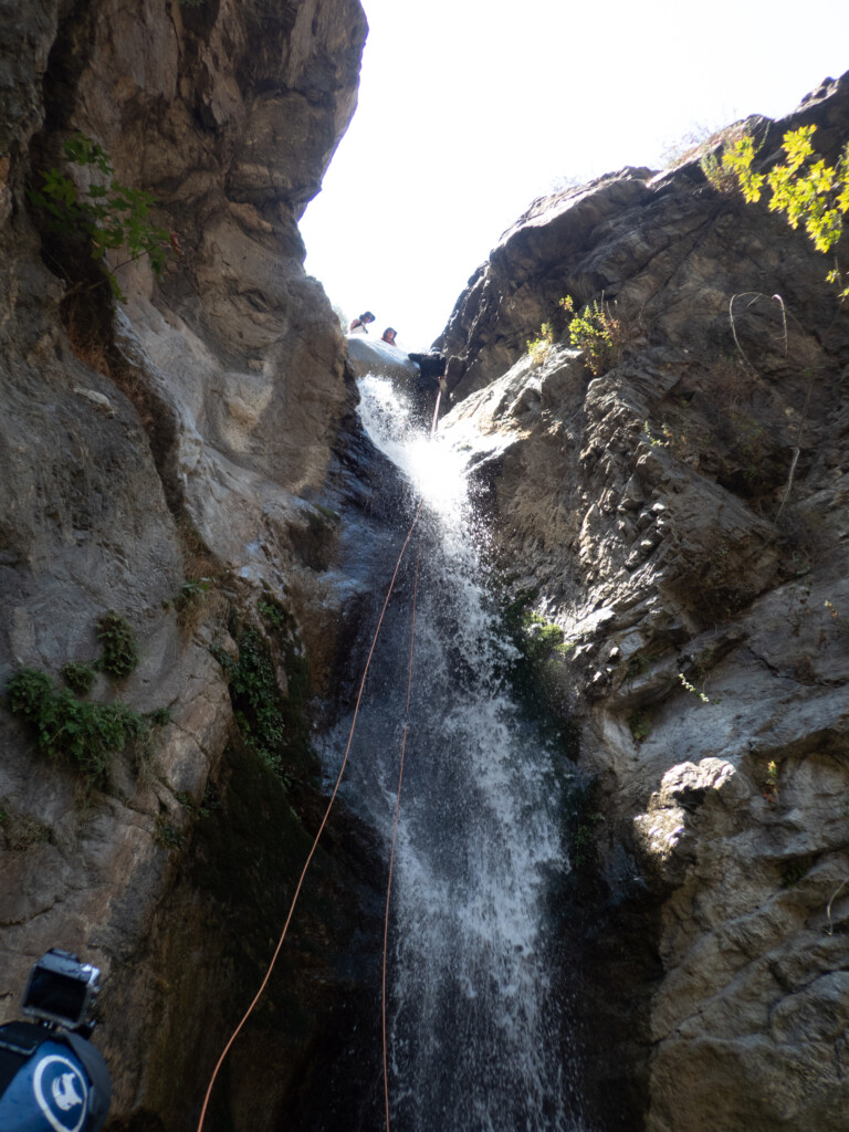 View looking up a rocky waterfall in Eaton Canyon where a person is rappelling down using ropes, water splashing around them, and two people observing from the top edge of the cliff under bright sky.