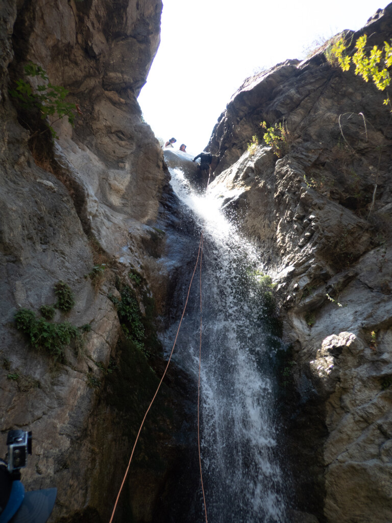 View looking up at a person rappelling down a waterfall in a rocky canyon with water splashing around them. Two people stand at the top of the waterfall watching. The canyon walls are steep with some green plants growing on the rocks. Bright sunlight illuminates the scene from above.
