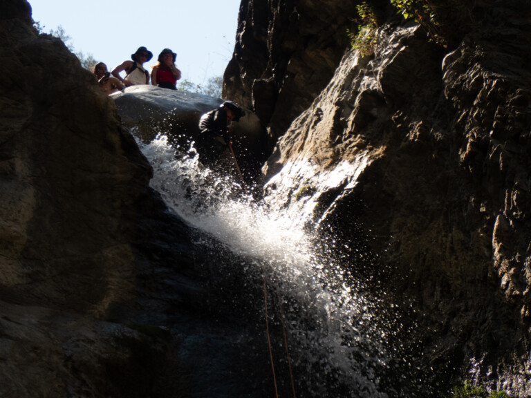 A person wearing a helmet and black clothing is rappelling down a waterfall in a rocky canyon, water splashing around them. Above, three people stand on a rock ledge watching, with sunlight illuminating parts of the canyon walls and water droplets.