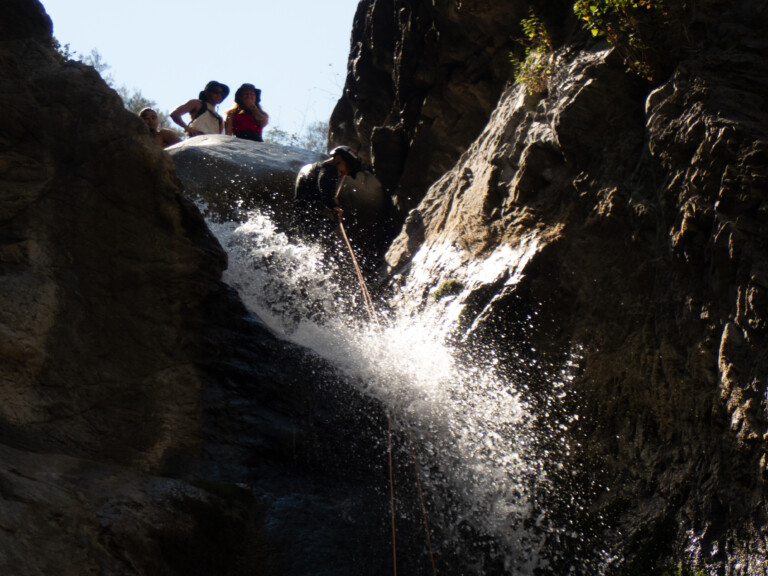 A person wearing a helmet and wetsuit rappelling down a waterfall in Eaton Canyon, with water splashing around. Three other people stand at the top of the waterfall, observing. The scene is surrounded by rocky canyon walls and some vegetation under clear blue sky.