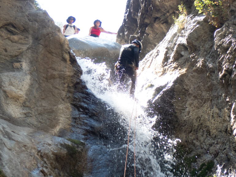 A person wearing a helmet and harness rappelling down a rocky waterfall in Eaton Canyon while two people wearing hats and backpacks watch from the top. Water splashes around the person descending, surrounded by steep rock walls and some vegetation.
