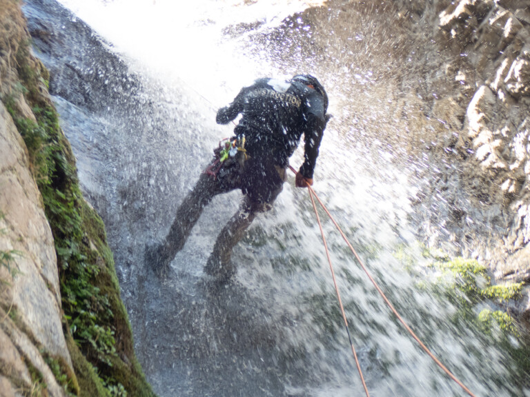 A person wearing a helmet and protective gear rappelling down a waterfall in Eaton Canyon, with water splashing around and rocky walls covered in moss and vegetation.