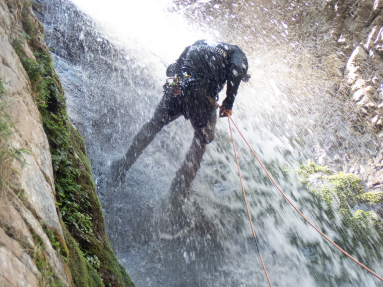 A person wearing a black jacket with the word 'CONSTRUCTION' on the back is rappelling down a waterfall at Eaton Canyon. Water sprays all around them as they descend using orange ropes, surrounded by rocky walls with patches of green moss and plants.
