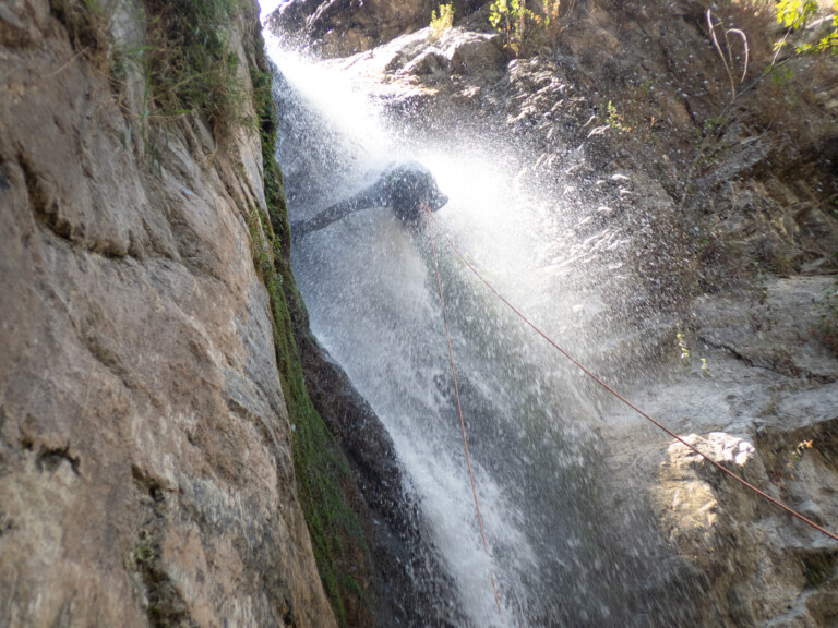 A person wearing a helmet and harness is rappelling down a waterfall in a narrow rocky canyon. Water sprays around them as they descend, with ropes securing their descent. The canyon walls are steep and covered with patches of moss and small plants.