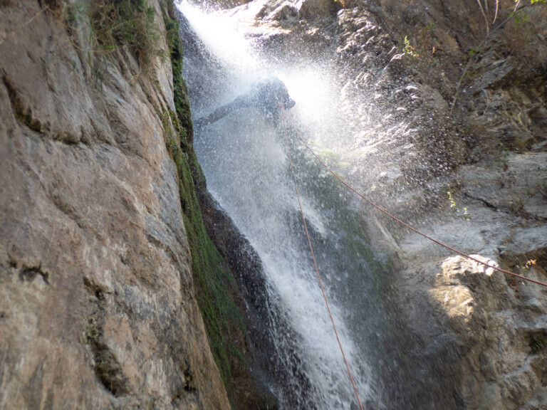 A person wearing a helmet and wetsuit is rappelling down a waterfall in a narrow rocky canyon. Water splashes heavily around them as they descend using ropes secured to the cliff. The canyon walls are steep with patches of moss and small plants.